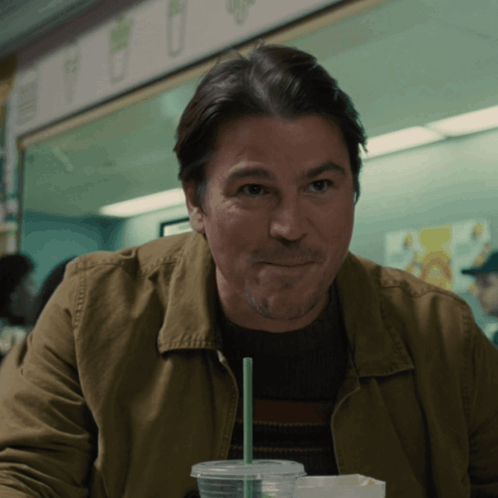 A Man In A Brown Jacket Is Sitting At A Table With A Cup Of Coffee And A Green Straw GIF