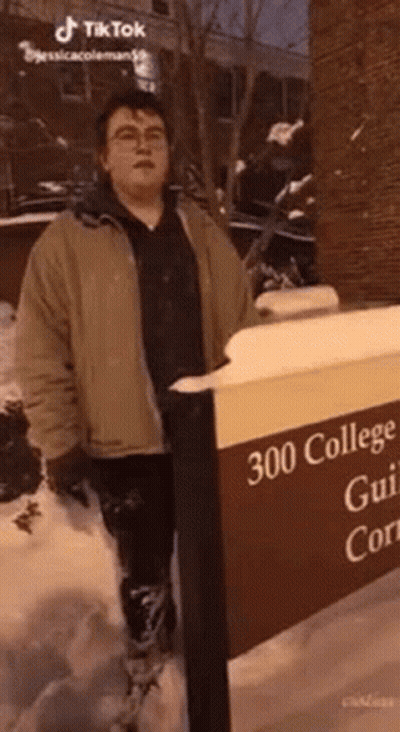 A Man In A Brown Jacket Is Standing Next To A Sign That Says `` 300 College Gui 'S Court '' . GIF