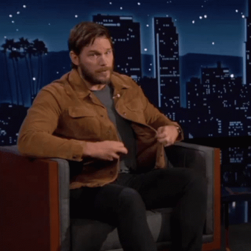 A Man In A Brown Jacket Sits In A Chair In Front Of A City Skyline GIF