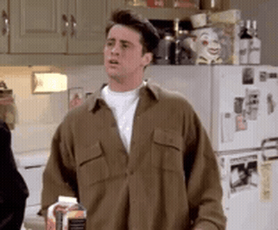 A Man In A Brown Shirt Is Standing In Front Of A Refrigerator Holding A Bottle Of Milk . GIF