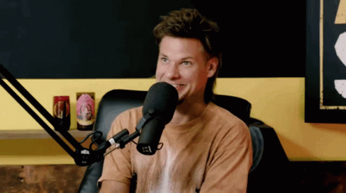 A Man In A Brown Shirt Is Talking Into A Microphone With A Can Of Beer On A Shelf Behind Him GIF