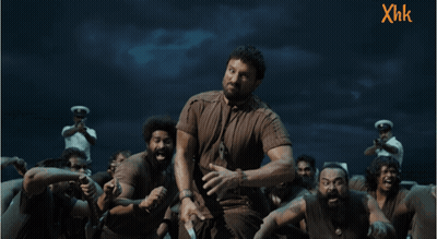 A Man In A Brown Shirt Stands In Front Of A Group Of People With Xhk Written On The Bottom GIF
