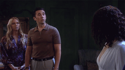 A Man In A Brown Shirt Stands Next To A Woman In A White Dress GIF