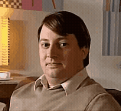 A Man In A Brown Sweater And White Shirt Is Sitting On A Couch Looking At The Camera . GIF