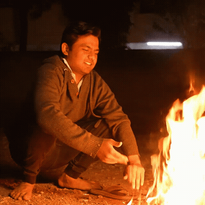 A Man In A Brown Sweater Is Kneeling Down Next To A Fire GIF