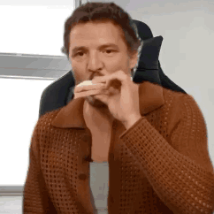 A Man In A Brown Sweater Is Sitting In A Chair Eating A Sandwich . GIF