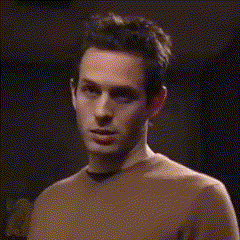 A Man In A Brown Sweater Is Standing In A Dark Room Looking At The Camera . GIF