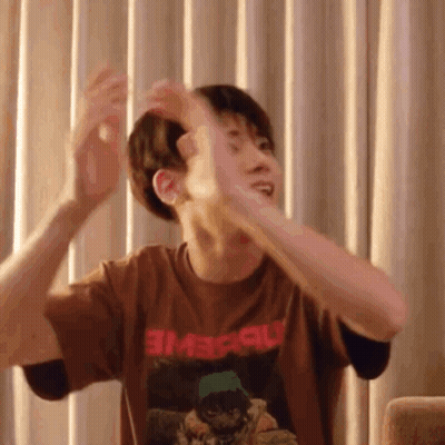 A Man In A Brown T-Shirt Is Sitting On A Couch With His Arms Outstretched In Front Of Curtains . GIF