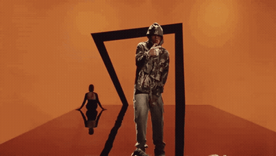 A Man In A Camo Hoodie Stands In Front Of A Black Triangle GIF