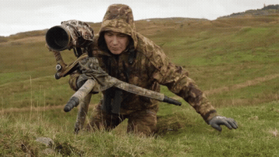 A Man In A Camo Jacket Is Kneeling Down In The Grass With A Camera On A Tripod GIF