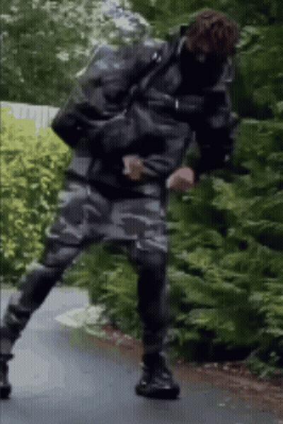 A Man In A Camouflage Jacket And Pants Is Standing On One Leg On A Road . GIF