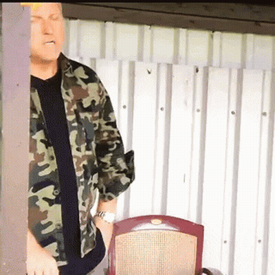A Man In A Camouflage Shirt Is Standing In Front Of A Chair GIF