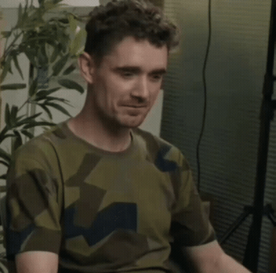A Man In A Camouflage Shirt Is Waving His Hand And Making A Funny Face . GIF