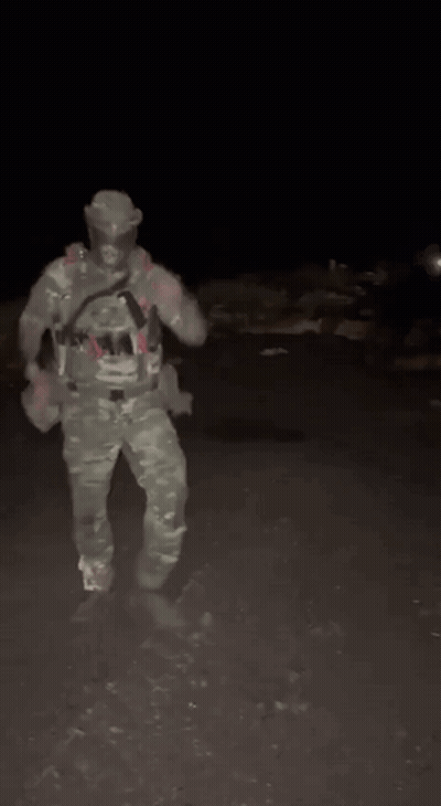 A Man In A Camouflage Uniform Is Standing In The Dark Holding A Gun . GIF