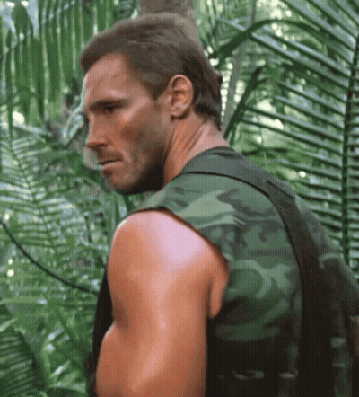 A Man In A Camouflage Vest Stands In A Jungle GIF