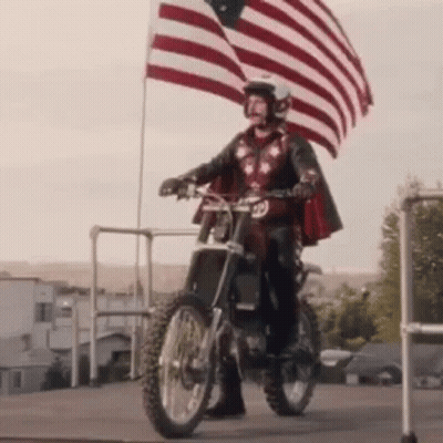 A Man In A Cape Is Riding A Dirt Bike While Holding An American Flag . GIF