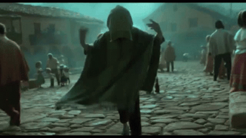 A Man In A Cape Is Walking Down A Cobblestone Street GIF