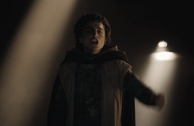 A Man In A Cape Stands In Front Of A Light In A Dark Room GIF