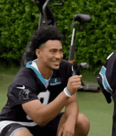 A Man In A Carolina Panthers Jersey Is Sitting On The Ground And Smiling . GIF