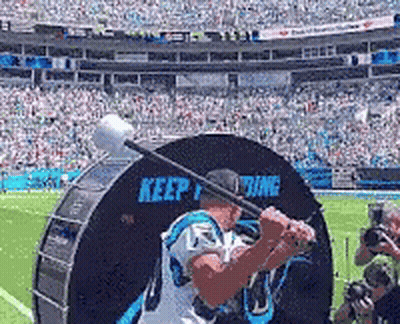 A Man In A Carolina Panthers Jersey Is Swinging A Bat At A Drum That Says Keep Fighting GIF