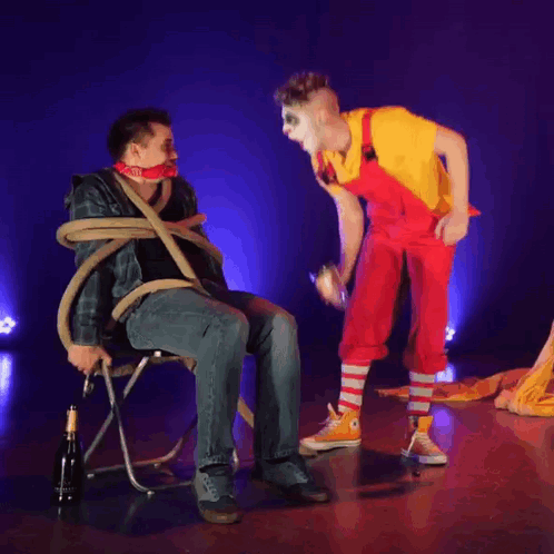 A Man In A Chair Is Tied Up By A Clown Who Is Holding A Bottle GIF