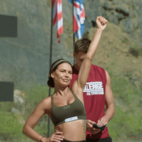 A Man In A Challenge Shirt Stands Next To A Woman In A Sports Bra GIF