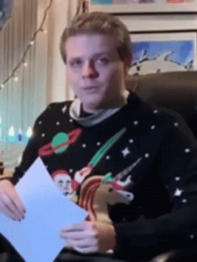 A Man In A Christmas Sweater Is Sitting In A Chair Holding A Piece Of Paper GIF