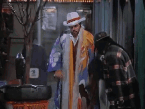 A Man In A Clown Costume Is Walking Down The Street With A Cane And A Hat . GIF