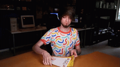 A Man In A Colorful Shirt Is Sitting At A Table Holding A Piece Of Paper That Says ' A ' On It GIF