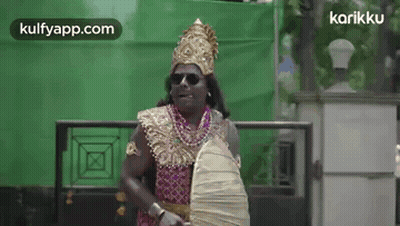 A Man In A Costume Is Standing In Front Of A Green Wall Holding A Fan And A Shield . GIF