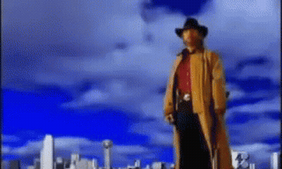 A Man In A Cowboy Hat And Trench Coat Stands In Front Of A City Skyline GIF