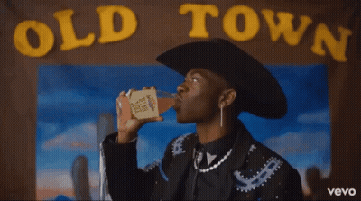 A Man In A Cowboy Hat Is Holding A Drink In Front Of An Old Town Poster GIF