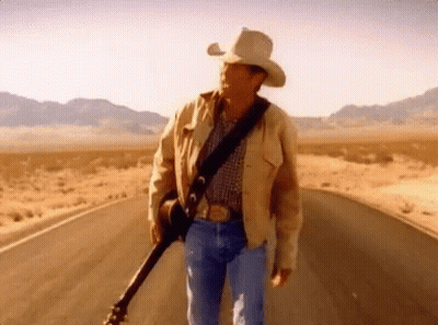 A Man In A Cowboy Hat Is Holding A Guitar While Walking Down A Road . GIF