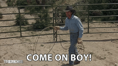 A Man In A Cowboy Hat Is Holding A Lasso In Front Of A Fence That Says Come On Boy GIF