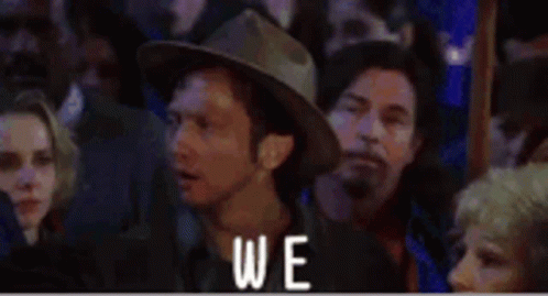 A Man In A Cowboy Hat Is Sitting In A Crowd Of People With The Word Again Written Above Him . GIF