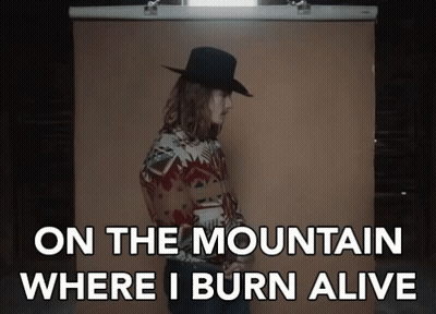 A Man In A Cowboy Hat Is Standing In Front Of A Brown Backdrop With The Words On The Mountain Where I Burn Alive GIF