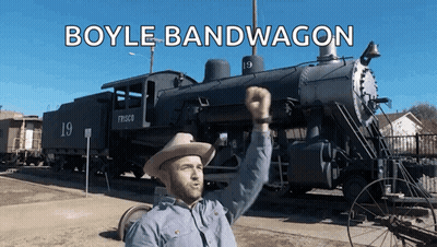 A Man In A Cowboy Hat Is Standing In Front Of A Train That Says Boyle Bandwagon On It GIF