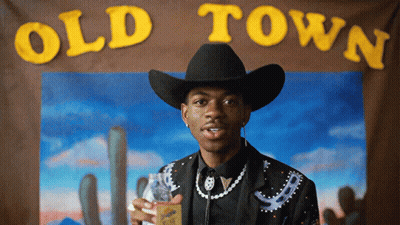 A Man In A Cowboy Hat Is Standing In Front Of An Old Town Sign GIF