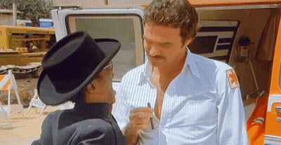 A Man In A Cowboy Hat Is Talking To Another Man In A White Shirt With A Red Cross On His Sleeve . GIF