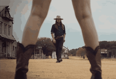 A Man In A Cowboy Hat Is Walking Down A Dirt Road Next To A Woman In Cowboy Boots GIF