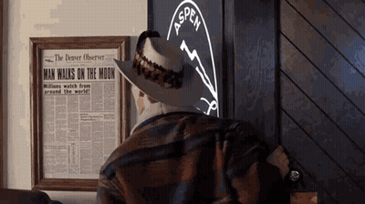 A Man In A Cowboy Hat Looks At A Framed Newspaper That Says Man Walks On The Moon GIF