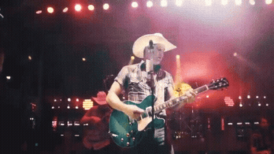 A Man In A Cowboy Hat Playing A Guitar On A Stage GIF