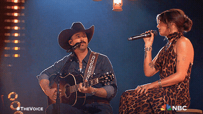 A Man In A Cowboy Hat Sings Into A Microphone While A Woman Sings Behind Him GIF