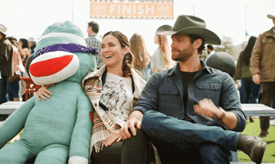 A Man In A Cowboy Hat Sits Next To A Woman Holding A Sock Monkey Under A Finish Sign GIF
