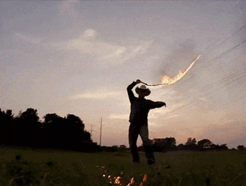 A Man In A Cowboy Hat Stands In A Field Holding A Flamethrower GIF
