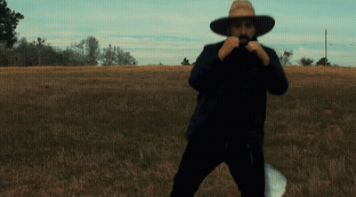 A Man In A Cowboy Hat Stands In A Grassy Field GIF