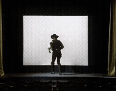 A Man In A Cowboy Hat Stands In Front Of A Large Screen GIF