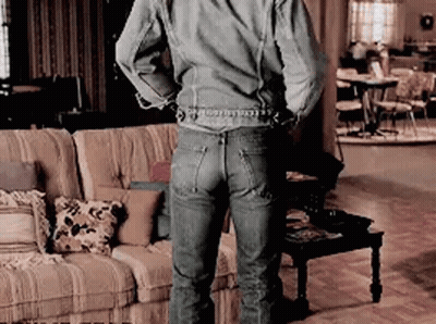 A Man In A Denim Jacket And Jeans Is Standing In A Living Room With His Hands Behind His Back . GIF