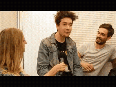A Man In A Denim Jacket Is Being Interviewed By A Woman While Two Other Men Sit Behind Him . GIF