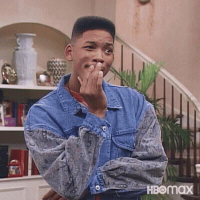 A Man In A Denim Jacket Is Covering His Mouth With His Hand While Standing In Front Of A Staircase . GIF